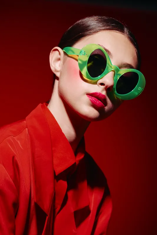 Fashion woman in green sunglasses with bright makeup and red lips poses against dark red background. Creative beauty look with vibrant colors and bold styling.