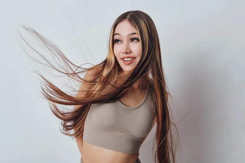 Young woman with long hair wearing casual tank top smiling and looking away on white background. Portrait of happy female with natural makeup and flowing hair in studio light.