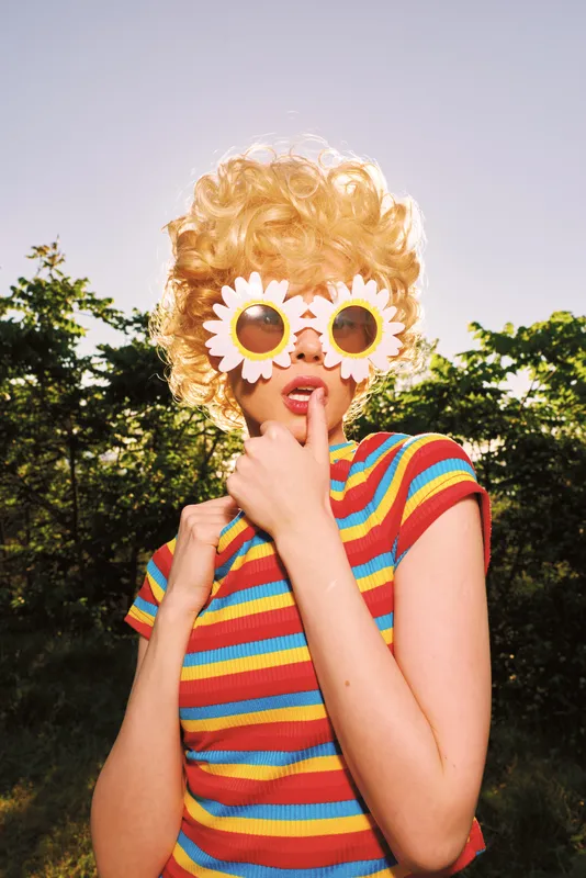 A playful woman in a colorful striped shirt outdoors wears flower sunglasses, creating a retro, cinematic vibe. Bright sun highlights vibrant hues and whimsical fashion.