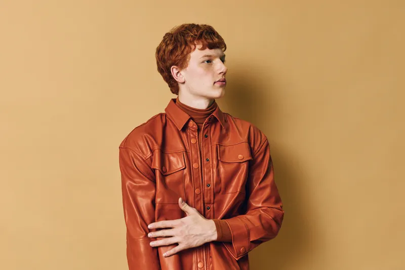 Young man with red hair wearing brown leather shirt posing in front of plain beige background. Fashion, casual style and portrait of confident male model in studio with neutral expression.