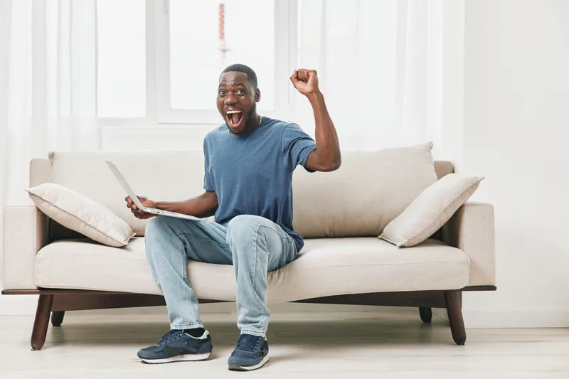 Excited young African American man celebrating success while sitting on a cozy sofa with a laptop in a bright, minimalistic living room