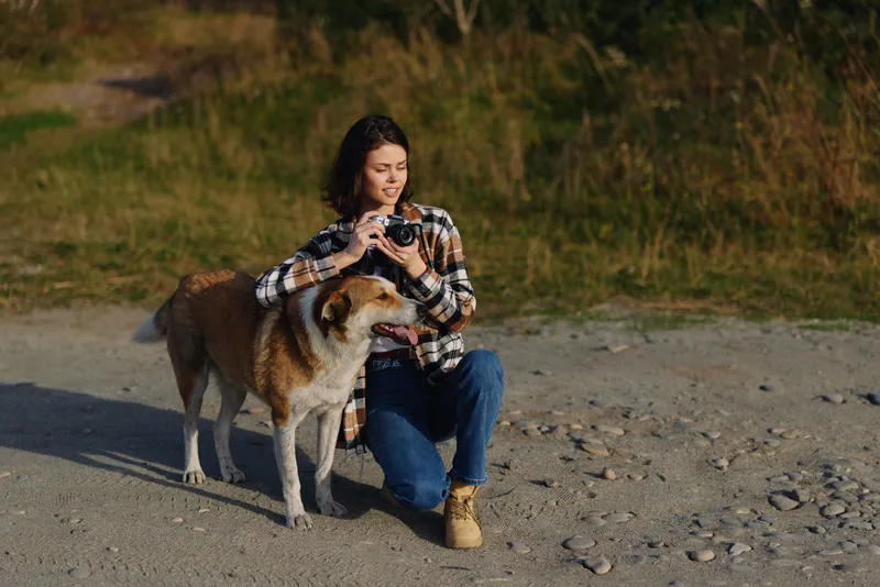 A photographer woman crouches beside a dog on a gravel path, capturing a candid moment with a compact camera in a natural outdoor setting among sparse vegetation on a sunny day.