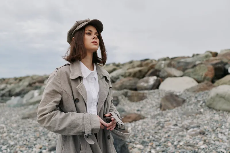 Young woman in vintage coat and cap stands on rocky beach, holding camera. Overcast sky creates moody atmosphere. Concept of retro style, travel, and outdoor photography.