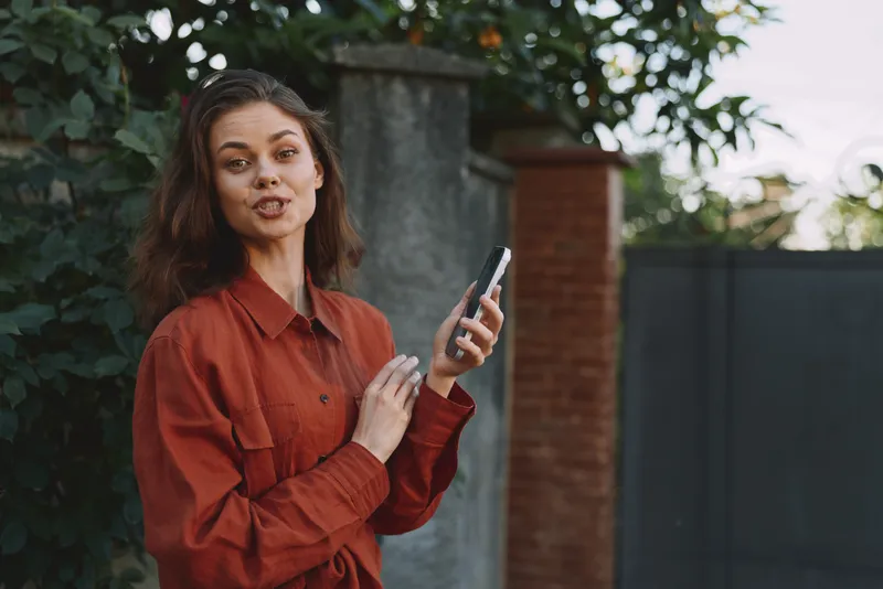 Smiling woman in a rust shirt checks her phone outdoors, urban garden backdrop. Focus on casual lifestyle, everyday technology use, relaxed mood, natural light