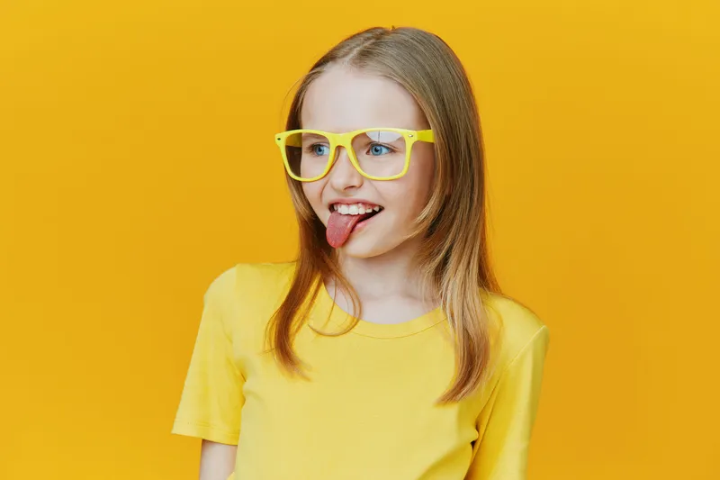Cheerful girl in yellow shirt and glasses showing playful tongue gesture against a vibrant yellow background, conveying joy and youthfulness