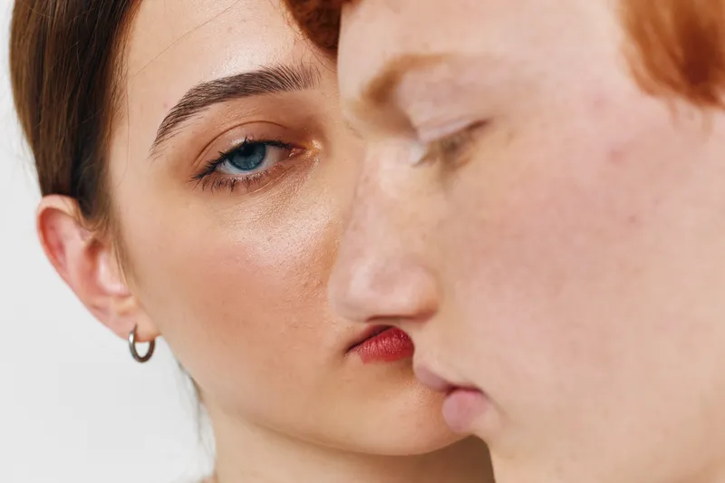 Two women close to each other with focus on the face, skin texture and natural makeup. Portrait of diverse girls with earrings and freckles on white background.