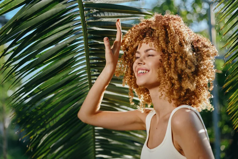 Smiling woman with curly hair enjoys sunlight outdoors near tropical palm leaves, wearing white tank top and radiating positive summer vibes in natural setting.