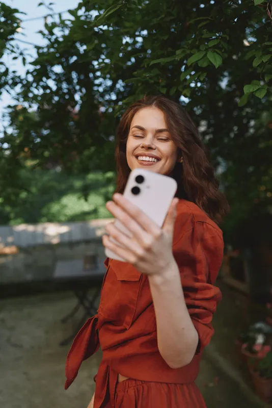 Vibrant lifestyle portrait of a smiling woman in a red dress taking a selfie outdoors, posing with her phone amid lush greenery and soft daylight ambiance