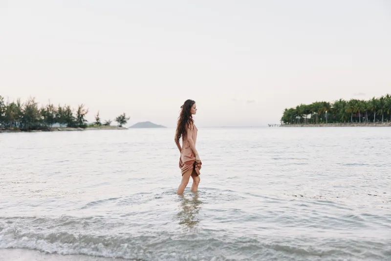 A solitary woman walks along the shoreline in shallow water, soft light, gentle waves, and a calm horizon, creating a serene coastal moment focused on solitude and nature.