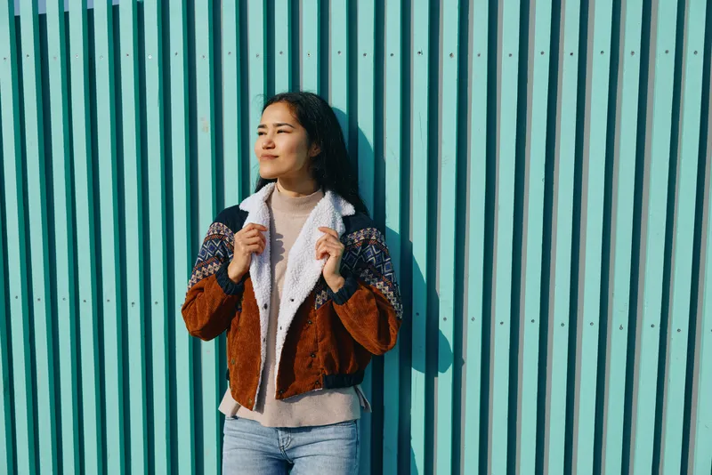 Casual fashion scene with a woman wearing a cozy jacket, denim jeans, and knit sweater, posing against a tall blue striped wall in urban outdoor setting during golden hour