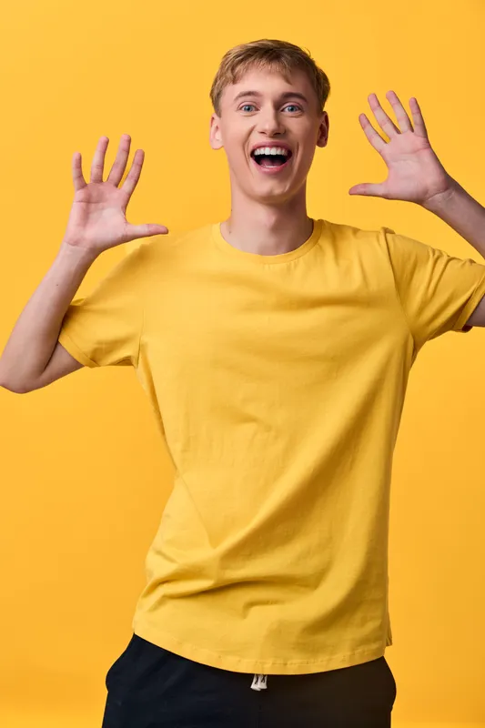 Excited young man with surprised expression, raised hands, yellow shirt and vibrant backdrop conveying happiness, energy and playful mood for lifestyle, fashion, and promotional visuals