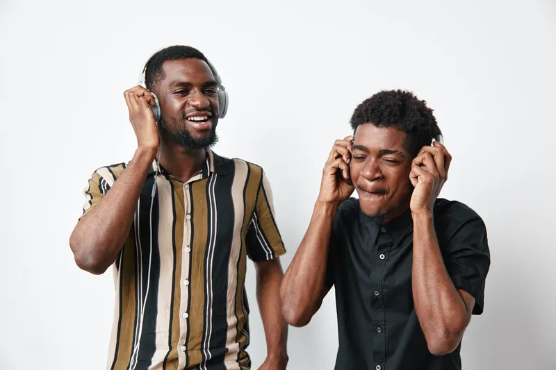 Two young black men with dark skin listening to music on headphones, enjoying sound and rhythm together in casual clothes with joyful expressions on white background.