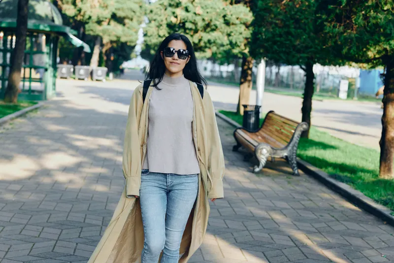 Woman walking in a park on a sunny day wearing a beige coat over a sweater, jeans, and sunglasses, showcasing casual outdoor fashion and street style against greenery and benches in a bright urban