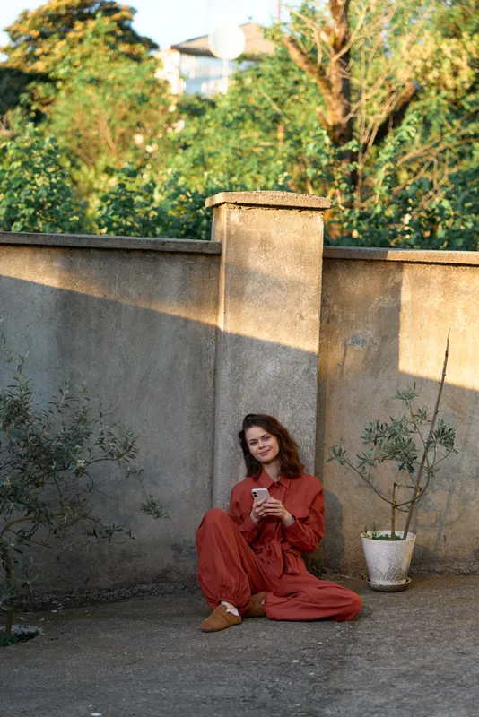 Young woman sits cross legged on the ground outdoors against a stone wall, wearing a red jumpsuit and holding a cup, enjoying a calm afternoon in golden light
