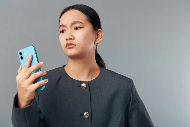 Woman holding smartphone with focused expression in studio setting. Young female dressed in dark outfit looking at phone screen thoughtfully on plain background for technology use.