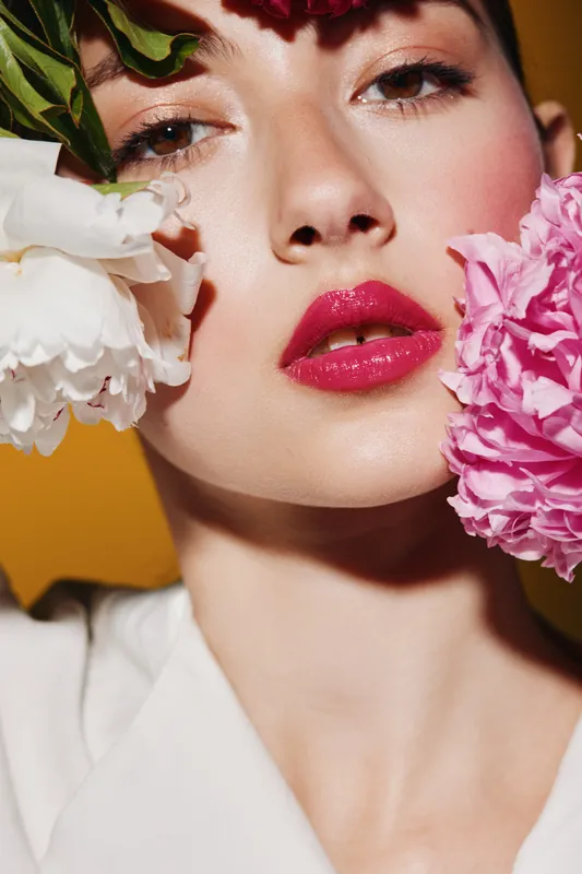 Close-up portrait of a young woman with creative beauty makeup and pink lipstick posing with white and pink peony flowers on yellow background. Fashion concept.