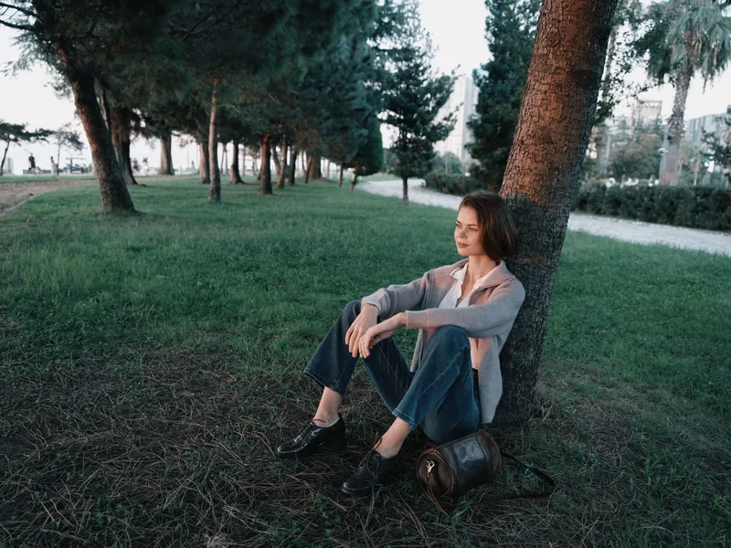 A thoughtful young professional sits on the grass beside a tree in a serene park setting, dressed in smart casual attire, appearing calm, focused, and reflective in natural light.