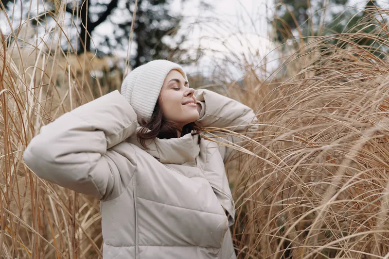 Woman in winter jacket outdoors among tall grass, serene smile, beanie and cozy coat for a calm natural portrait in chilly daylight