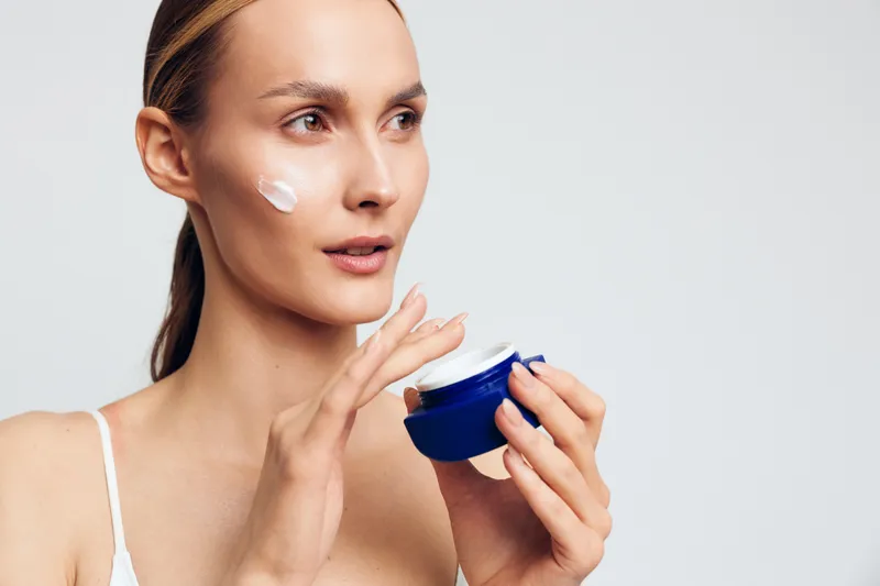 Young woman with clear skin applying white face cream on cheek holding blue jar. Skincare routine concept with natural beauty and self-care on white background. Soft natural lighting.