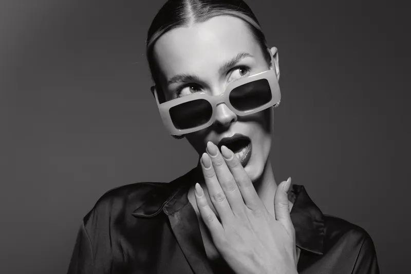 Young white woman with pale skin wearing stylish sunglasses and black satin blouse looks surprised covering mouth with hand in studio. Fashion and beauty portrait concept.