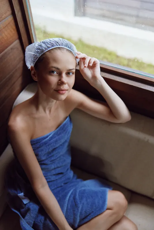 Young woman wrapped in a blue towel wearing a shower cap sitting by a window, relaxing indoors with natural light enhancing her calm expression and casual posture.