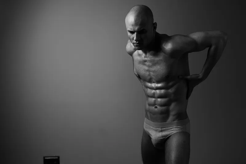 Black and white image of a muscular man showcasing his physique, emphasizing strength and determination in a minimalist setting.