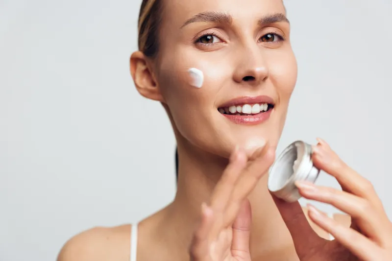 Young woman with smooth skin applies white facial cream on her cheek in a bright studio, smiling softly as soft light highlights a healthy glow.