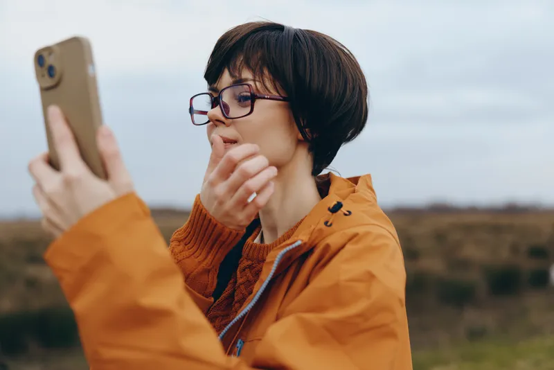 Woman wearing orange jacket and glasses outdoors holds a smartphone close, casual thinking expression, lifestyle communication portrait in open landscape and field bright sky during windy day