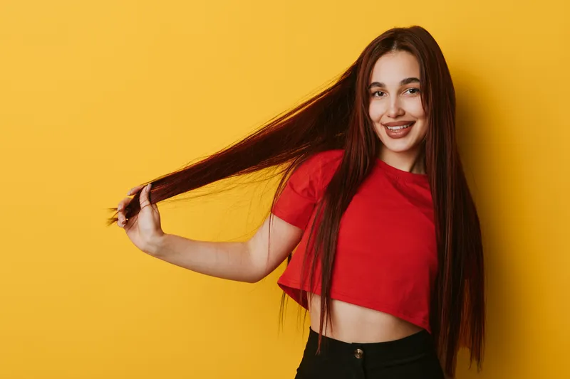 Young woman with long straight hair wearing red crop top and black pants smiling and holding hair on yellow background. Female model posing with casual style and happy expression.