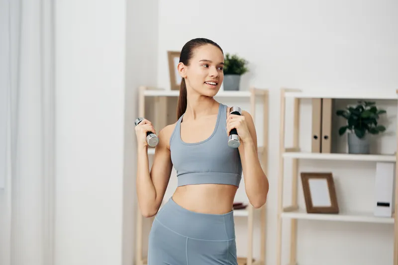 Woman fitness sport in a home gym lifts dumbbells, wearing grey sportswear. Bright room with shelves, plant, and decor; focused on strength training, posture, and healthy lifestyle.