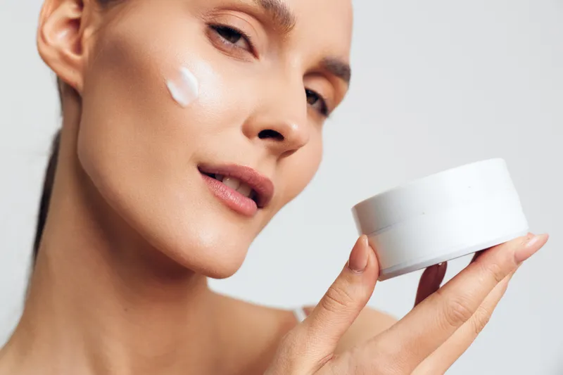 A woman applies white cream on her cheek while holding a white cosmetic jar. The close-up shot highlights skincare routine and facial care with a clean background.