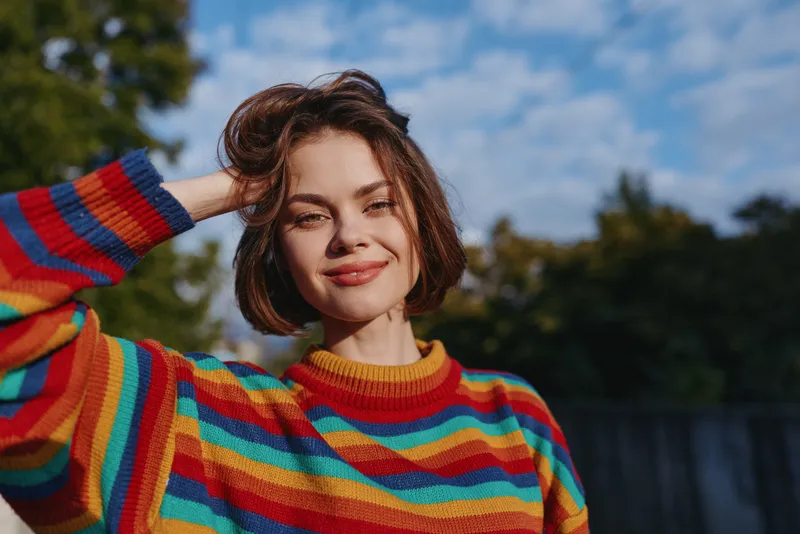Woman smiling in colorful striped sweater, short hair portrait outdoors in warm sunlight. Young adult model in casual outfit, hand on head, relaxed pose and natural confident lifestyle portrait.