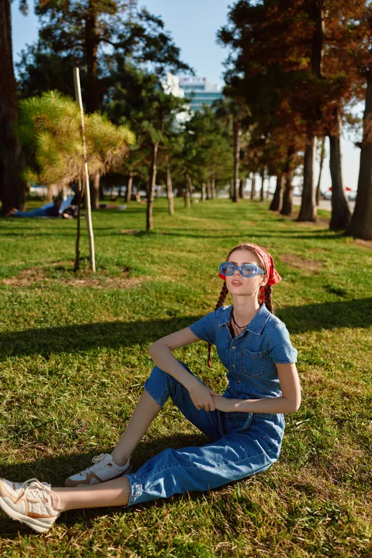 young woman sitting on grass in park wearing denim outfit and sunglasses enjoying sunny day with trees and green lawn in background
