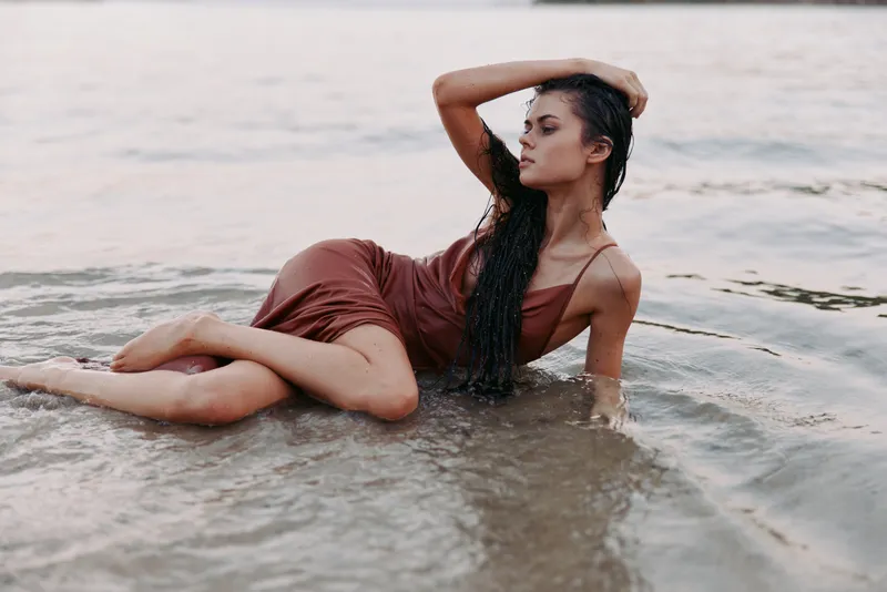 Elegant woman lounges in shallow water wearing a warm-toned dress, relaxed pose, and wet hair, creating a serene moment with soft light and calm shoreline ambiance.
