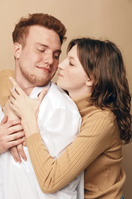 Young couple embraces tenderly with eyes closed, expressing love and affection. Woman in beige sweater hugs man in white shirt against neutral background. Concept of romance and intimacy.
