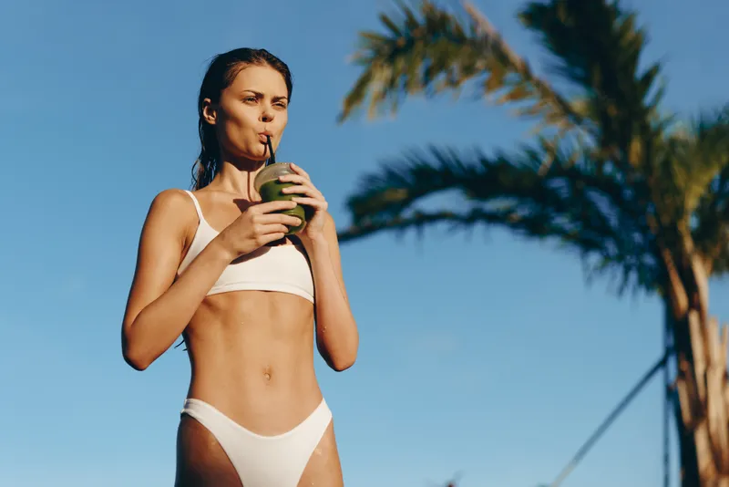 A young woman in a white bikini sips from a coconut on a sunny beach, her relaxed posture and distant gaze capturing a carefree vacation moment under clear blue skies.