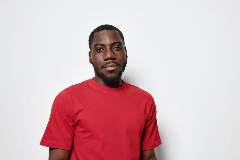 African American man with dark skin wearing red t shirt standing against white background looking at camera with calm expression and relaxed posture.