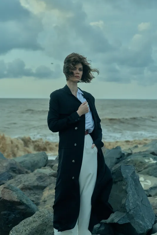 Woman in a long dark coat over a white shirt stands on a rocky coast, wind swirling her hair, a moody seascape and dramatic sky highlighting modern fashion on the shore