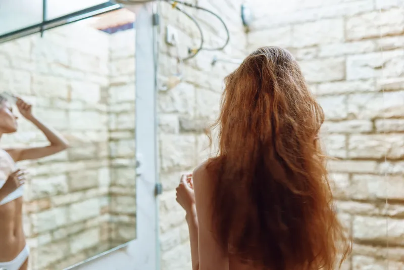 Woman with long hair standing before bathroom mirror with stone wall background, natural light, self-care routine, serene atmosphere, morning glow.