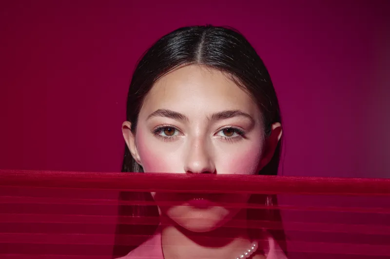 Young woman in pink makeup stands behind translucent red fabric, revealing intense gaze. Studio shot with deep maroon background. Concept of beauty, fashion, and creative skincare.