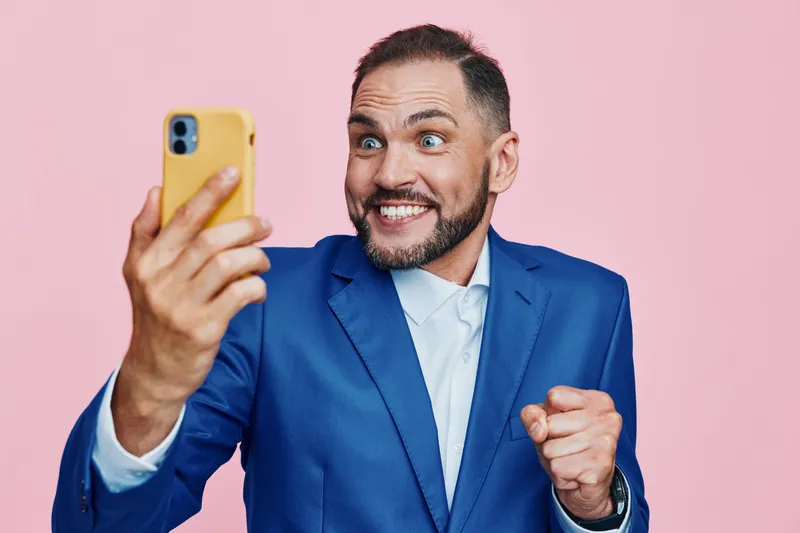A professional businessman on a solid pink background takes a selfie with a lively, surprised expression, projecting energy, confidence, and dynamic leadership.