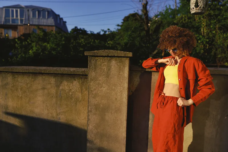 Young woman in a bright orange jacket and pants leans against a concrete wall in sunlight, striking a confident street style pose, her cropped yellow top peeking out and the urban setting emphasizing