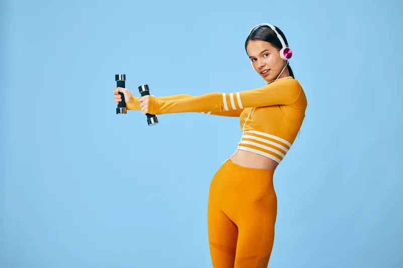 Young woman fitness model in bright orange activewear holds small dumbbells while stretching arms, wearing pink headphones against a clean blue studio background.
