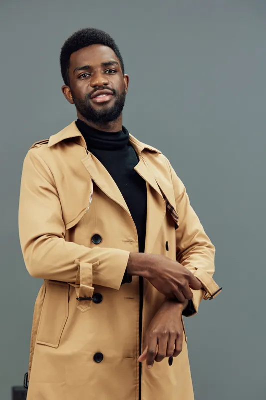 Stylish African man in a beige trench coat expressing confidence and style against a muted background, perfect for fashion concepts and seasonal promotions