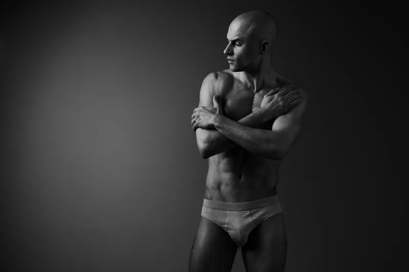 Black and white portrait of a man in a pose, conveying strength and introspection, showcasing his muscular physique and thoughtful expression.