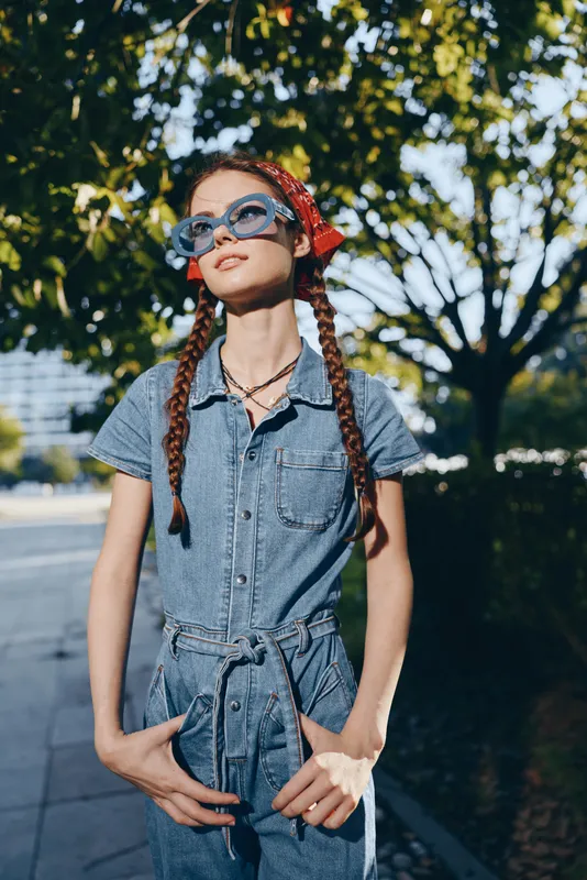 Lifestyle woman in modern boho-western denim street style wearing sunglasses, with braided hair, outdoors under vibrant green trees, film color effect.
