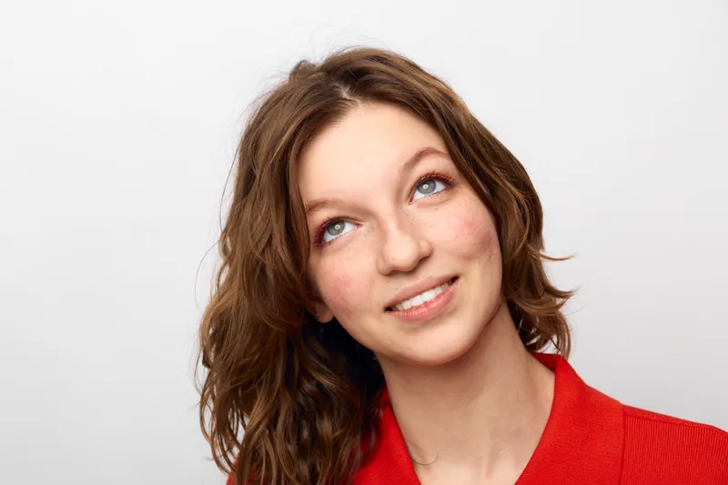 Young woman with brown hair and blue eyes smiles gently looking up. She wears a red shirt against a plain white background. Concept of hope, optimism, and casual portrait.