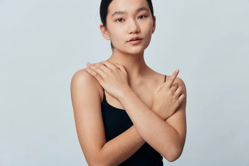 Young woman with natural skin in black top posing against light background crossing arms on chest, looking calm and confident with minimal makeup and simple hairstyle.