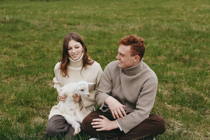 Young couple sitting on grass with a baby goat in their arms enjoying a relaxed lifestyle moment in cozy sweaters during a calm outdoor day.