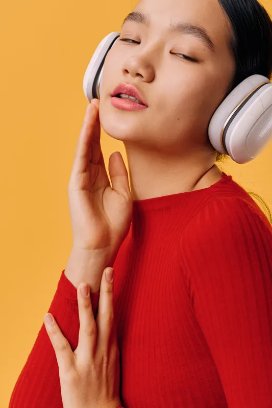 Young woman in red sweater wearing white headphones with closed eyes listening to music, feeling relaxed and enjoying sound, isolated on orange background with hand near face.